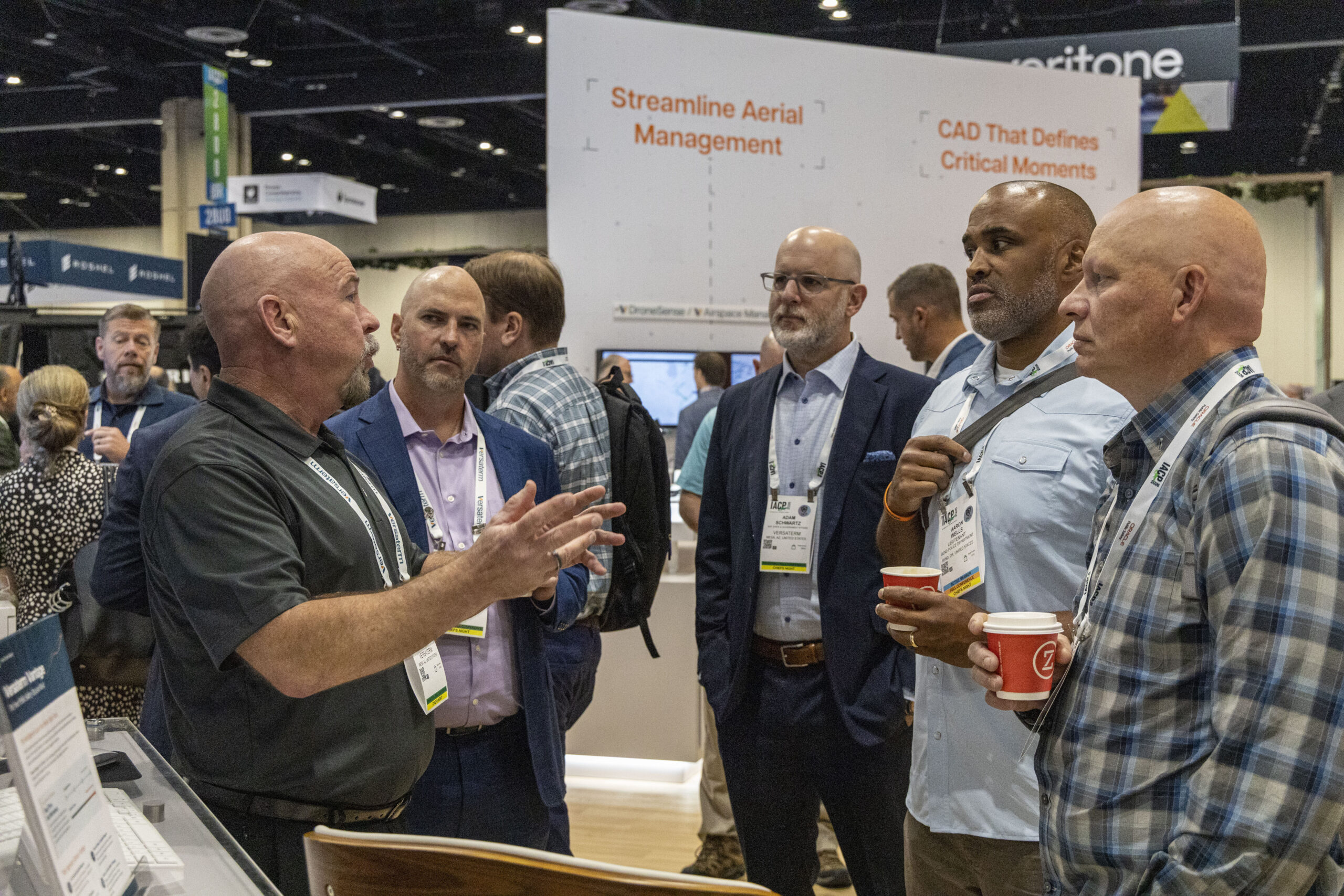 A group of attendees at the Versaterm booth at IACP, engaged in conversation near signage reading "Streamline Aerial Management" and "CAD That Defines Critical Moments."